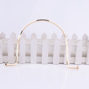 May include: Gold metal handbag handle with a rounded top and two curved ends. The handle is designed to be attached to a handbag.