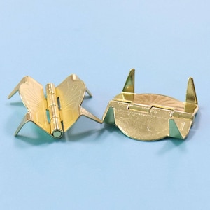 May include: Two gold-colored butterfly-shaped hinges. One hinge is fully open, displaying the intricate details of the butterfly wing design. The other hinge is partially closed. These decorative hinges are ideal for small craft projects.