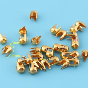 May include: A collection of small, gold-colored metal studs with a three-pronged design. The studs are approximately 0.12 inches wide and 0.16 inches tall. They are scattered on a light blue surface, suitable for various craft and design projects.