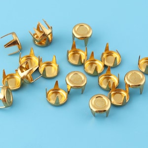 May include: A collection of gold-toned metal studs with four prongs. The studs are arranged on a light blue background.