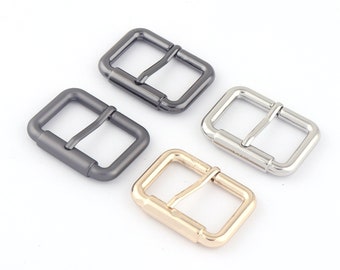 May include: Four metal belt buckles in different finishes: silver, gold, black, and gunmetal. Each buckle has a rectangular shape with rounded corners and a center bar for securing a belt.