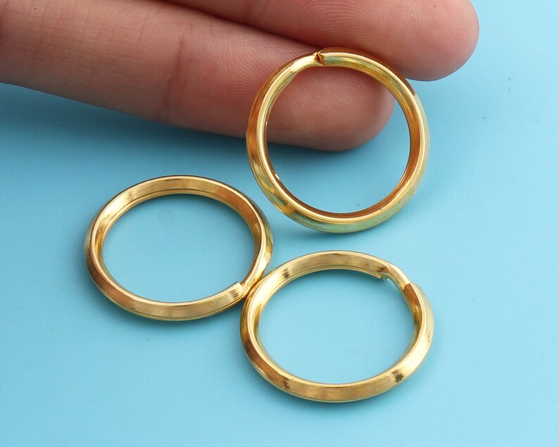 May include: Three gold-colored key rings are displayed against a light blue background. The rings are circular with a small opening, suggesting they are designed to hold keys or other small items. The rings have a smooth, metallic finish.