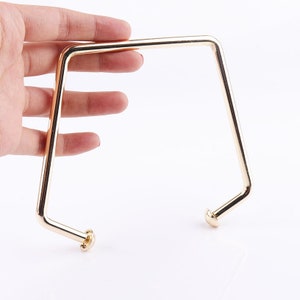 May include: Gold-toned metal handbag handle with rounded ends. The handle is shaped like a rectangle with rounded corners.