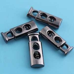 Puede incluir: Cuatro cierres de palanca de metal negro con dos agujeros cada uno. Los cierres tienen un mecanismo de resorte y se utilizan para asegurar cordones o correas.