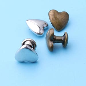 K&ouml;nnte beinhalten: Vier herzf&ouml;rmige Metallkn&ouml;pfe, zwei silberne und zwei bronzene. Die silbernen Kn&ouml;pfe sind flach, w&auml;hrend die bronzenen Kn&ouml;pfe einen Stift auf der R&uuml;ckseite zum N&auml;hen haben.