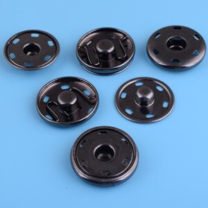 May include: Six black metal snap buttons with a shiny finish. The buttons have a round shape with a raised center and a series of small holes around the edge.