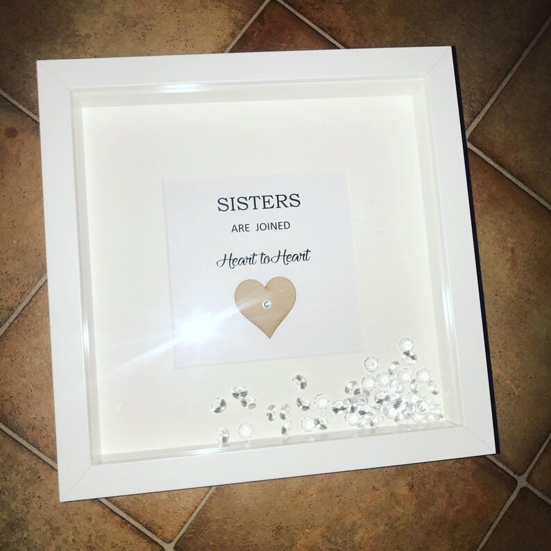 Op de afbeelding: Witte fotolijst met een witte passe-partout en een heldere glazen voorkant. De lijst bevat een kaart met de tekst "Sisters are joined Heart to Heart" en een houten hartje met een strasssteen in het midden. De lijst is gevuld met heldere strassstenen.
