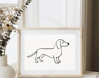 May include: A framed print with a minimalist black line drawing of a dachshund dog. The artwork is set against a cream background and framed in light wood. The print is displayed on a white surface near a window.