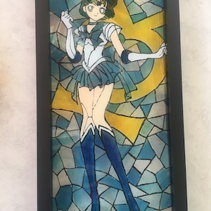 May include: A stained glass framed artwork featuring a cartoon character with green hair and a blue and white outfit. The character is standing in front of a yellow and blue background.