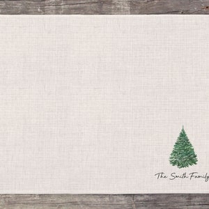 May include: A white linen placemat with a green watercolor illustration of a Christmas tree and the text "The Smith Family".