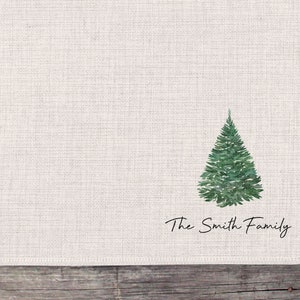 May include: A white linen napkin with a green watercolor Christmas tree and the text "The Smith Family" printed in black.