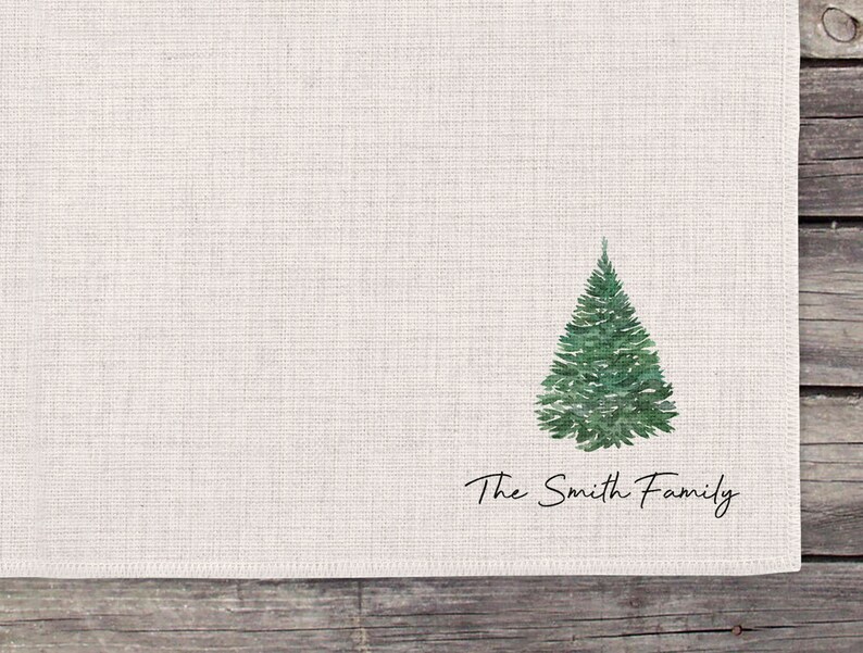 May include: A white linen napkin with a green watercolor Christmas tree and the text "The Smith Family" printed in black.