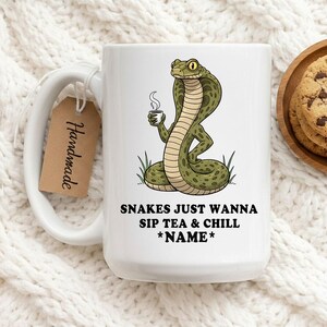 Puede incluir: Taza de cerámica blanca con una ilustración de una cobra de dibujos animados sosteniendo una taza de té. La taza tiene el texto "Snakes Just Wanna Sip Tea & Chill *Name*". Una etiqueta marrón que dice "Handmade" está adjunta al asa.