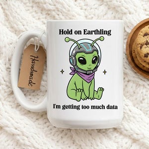 Op de afbeelding: Witte keramische mok met een groene alien in een ruimtehelm en paarse sjaal. Tekst op de mok luidt: "Hold on Earthling, I'm getting too much data." Een "Handmade" label is bevestigd. Op een witte gebreide ondergrond.