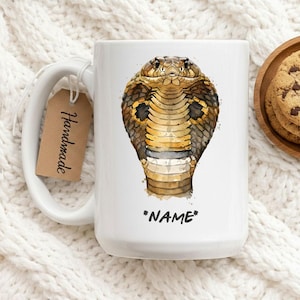 Puede incluir: Taza de cerámica blanca con una ilustración de acuarela de una cobra. La serpiente está representada en tonos marrones, dorados y negros. La taza tiene la palabra "*NAME*" impresa debajo. Una etiqueta que dice "Handmade" está unida al asa.