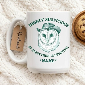May include: White ceramic mug with a humorous owl design. The mug features a sketch of an owl wearing a cowboy hat, with the text "HIGHLY SUSPICIOUS OF EVERYTHING & EVERYONE" in green. A small tag that says "Handmade" is attached.