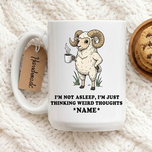 May include: White ceramic mug with a humorous illustration of a sheep holding a coffee cup. The mug features the text "I'M NOT ASLEEP, I'M JUST THINKING WEIRD THOUGHTS *NAME*". A "Handmade" tag hangs from the handle.