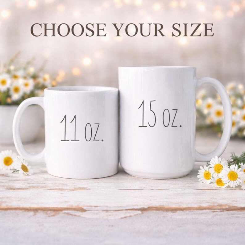 Op de afbeelding: Twee witte keramische mokken, &eacute;&eacute;n met het opschrift "11 oz." en de andere met "15 oz.". De tekst "CHOOSE YOUR SIZE" staat boven de mokken. De mokken staan op een wit houten oppervlak met madeliefjes.