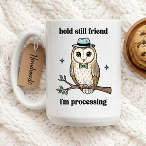 May include: White ceramic mug with a cartoon owl wearing a blue hat and bow tie, perched on a branch. The mug has the text "hold still friend" and "i'm processing". A brown tag that says "Handmade" is attached to the handle.