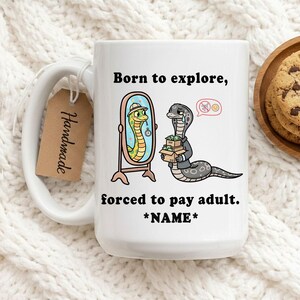 Puede incluir: Taza de cerámica blanca con una ilustración humorística de una serpiente. La taza presenta una serpiente de dibujos animados mirándose en un espejo y otra serpiente que lleva cajas de dinero. El texto dice "Born to explore, forced to pay adult. *NAME*". Una etiqueta "Handmade" cuelga del asa.