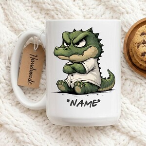May include: White ceramic mug featuring a cartoon alligator with an angry expression, wearing a white shirt. The mug has the word "*NAME*" printed on it. A brown tag with the word "Handmade" hangs from the handle.