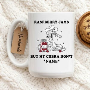Op de afbeelding: Witte keramische mok met een humoristische afbeelding van een cobra met een cowboyhoed en een pistool, met de tekst "RASPBERRY JAMS BUT MY COBRA DON'T *NAME*". Inclusief een "Handmade" label.