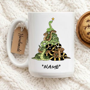 May include: White ceramic mug with a Christmas tree design made of cartoon snakes in green, brown, and black. The mug has a handle and a tag that says "Handmade". The text "*NAME*" is at the bottom.