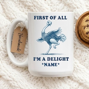 Può includere: Tazza in ceramica bianca con un manico grande, con un'illustrazione blu di uno struzzo e il testo "FIRST OF ALL I'M A DELIGHT *NAME*". Una piccola etichetta marrone con la scritta "Handmade" è attaccata al manico.