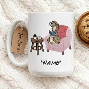 May include: White ceramic mug featuring a cartoon snake reading a book while seated in a pink armchair. A small wooden table with a plant and a cup is next to the chair. The mug has the word "*NAME*" and a "Handmade" tag.