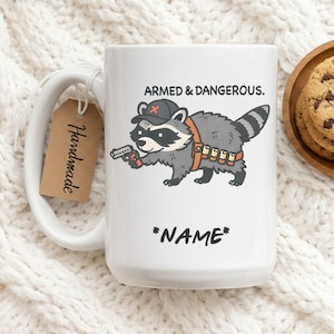 Peut inclure: Mug en céramique blanche avec un dessin de raton laveur portant un chapeau et tenant une arme à feu. Le texte "ARMED & DANGEROUS." est au-dessus. Le texte "*NAME*" est en dessous. Une étiquette "Handmade" est attachée.
