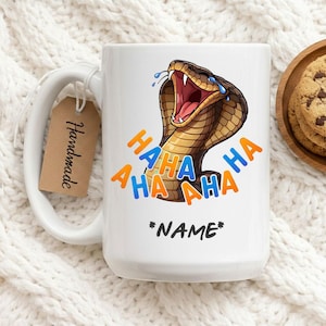 Puede incluir: Taza de cerámica blanca con un diseño de cobra de dibujos animados, con la boca abierta y rodeada de texto "HAHA" en naranja y azul. La taza tiene una etiqueta que dice "Handmade" y espacio para un nombre.