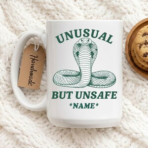 Peut inclure: Mug en céramique blanche avec une illustration de cobra verte et le texte "UNUSUAL BUT UNSAFE *NAME*". Une étiquette marron avec "Handmade" est attachée. Le mug est posé sur une surface tricotée blanche.