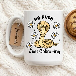 May include: White ceramic mug with a large handle, featuring a cartoon cobra illustration with daisies and the text "NO RUSH Just Cobra-ing". A handmade tag hangs from the handle. A wooden plate with cookies is in the background.