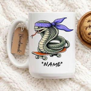 Pode incluir: Caneca de cerâmica branca com uma cobra de desenho animado usando uma bandana roxa e andando de skate laranja. A cobra tem uma língua vermelha. A caneca tem a palavra "*NAME*" impressa.
