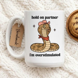 Könnte beinhalten: Weiße Keramik-Tasse mit einer humorvollen Cartoon-Schlange, die einen rosa Cowboyhut und ein rotes Halstuch trägt. Auf der Tasse steht der Text "hold on partner" und "I'm overstimulated". Ein "Handmade"-Etikett hängt am Griff.