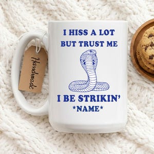May include: White ceramic mug with a blue cobra illustration and the text "I HISS A LOT BUT TRUST ME I BE STRIKIN' *NAME*". A brown tag that says "Handmade" is attached to the handle. Chocolate chip cookies are on a wooden plate.