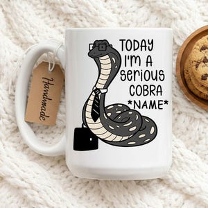 Op de afbeelding: Witte keramische mok met een cartoon cobra met een bril, stropdas en aktetas. De tekst op de mok luidt "TODAY I'M A serious COBRA *NAME*". Een bruin label met "Handmade" is bevestigd.