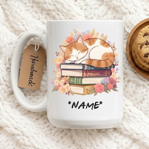 May include: White ceramic mug featuring a sleeping orange and white cat atop a stack of books, surrounded by flowers. The mug has a large handle and a tag that says "Handmade". The word "*NAME*" is printed on the mug.