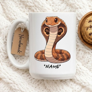 Puede incluir: Taza de cerámica blanca con una ilustración de una cobra de dibujos animados en tonos marrón, beige y negro. La cobra tiene su capucha extendida. El texto "*NAME*" está debajo de la cobra. Una etiqueta marrón con la palabra "Handmade" cuelga del asa.