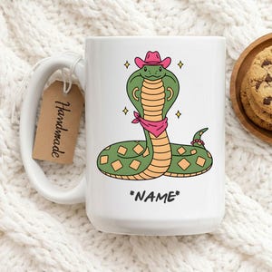 May include: White ceramic mug featuring a cartoon cobra wearing a pink cowboy hat and bandana. The snake is green and orange with diamond patterns. The mug has the text "*NAME*" and a tag that says "Handmade".