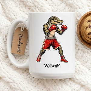 Könnte beinhalten: Weiße Keramik-Tasse mit einem Cartoon-Alligator-Boxer in roten Shorts, Handschuhen und Schuhen. Der Alligator trägt ein weißes Tanktop. Die Tasse hat ein Etikett mit der Aufschrift "Handmade" und das Wort "NAME" ist auf die Tasse gedruckt.