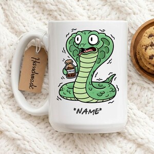 Op de afbeelding: Witte keramische mok met een cartoon cobra ontwerp. De groene cobra houdt een espressofles vast en lijkt te zweten. De mok heeft een label met de tekst "Handmade" en de tekst "*NAME*".