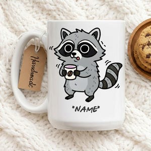 Könnte beinhalten: Weiße Keramik-Tasse mit einem Cartoon-Waschbären, der eine Kaffeetasse hält. Der Waschbär ist grau und schwarz mit einem gestreiften Schwanz. Die Tasse hat den Text "*NAME*" und ein "Handmade"-Etikett.