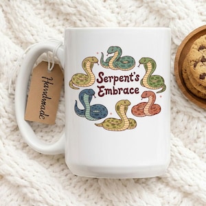 May include: White ceramic mug with a colourful illustration of six cartoon snakes around the text "Serpent's Embrace." The snakes are in shades of green, blue, yellow, and red. A brown tag with the word "Handmade" hangs from the handle.