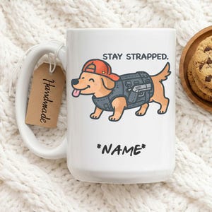 Peut inclure: Mug en céramique blanche avec un chien de dessin animé portant un gilet tactique et une casquette rouge. Le mug porte le texte "STAY STRAPPED." au-dessus du chien et "*NAME*" en dessous. Une étiquette faite à la main est attachée à la poignée.