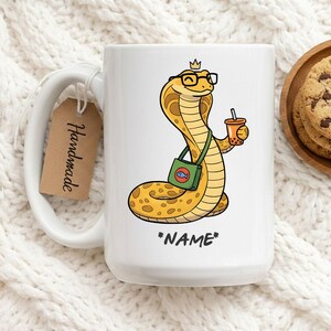 Puede incluir: Taza de cerámica blanca con una cobra de dibujos animados con gafas y una corona, sosteniendo un té de burbujas y una bolsa verde con un emblema de pez. La taza tiene el texto "*NAME*" y una etiqueta "Handmade".