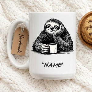 May include: White ceramic mug featuring a black and white illustration of a sloth holding a coffee cup. The sloth is resting its head on its hand. The mug has the word "*NAME*" printed on it. A brown tag that says "Handmade" is attached to the mug.