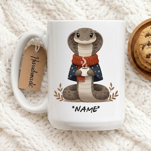 May include: White ceramic mug with a cartoon cobra design. The cobra wears a red scarf and blue cape, holding a coffee cup. The mug has the text "*NAME*" at the bottom and a "Handmade" tag. Cookies are on a wooden plate in the background.