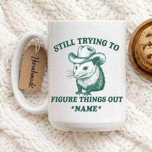 May include: White ceramic mug with a green illustration of an opossum wearing a cowboy hat. The mug has the text "STILL TRYING TO FIGURE THINGS OUT *NAME*". A brown tag that says "Handmade" is attached to the handle.