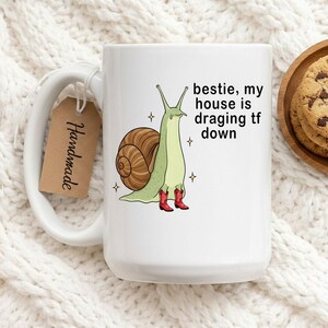 Op de afbeelding: Witte keramische mok met een cartoon slak met rode cowboylaarzen. De slak heeft een bruin huisje en is omringd door sterren. De tekst op de mok luidt "bestie, my house is dragging tf down". Een "Handmade" label hangt aan het handvat.